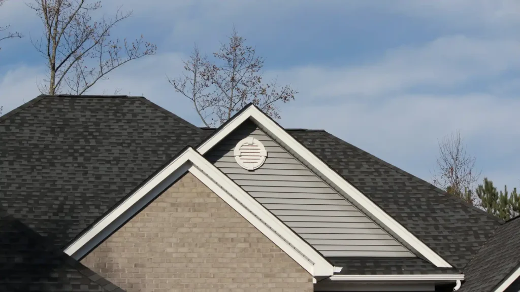 A roof with dark shingles and a vent installed on the gable end of the house, surrounded by siding and a clear sky in the background.
