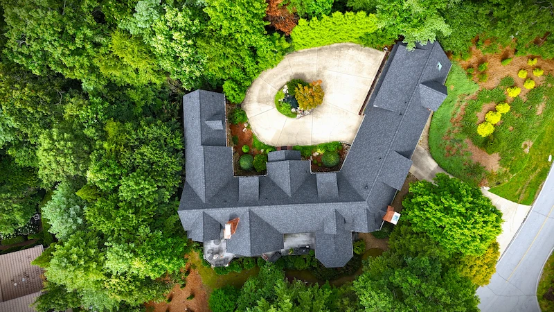 Direct overhead aerial view of a dark grey architectural shingle roof on a large L-shaped house surrounded by dense trees.